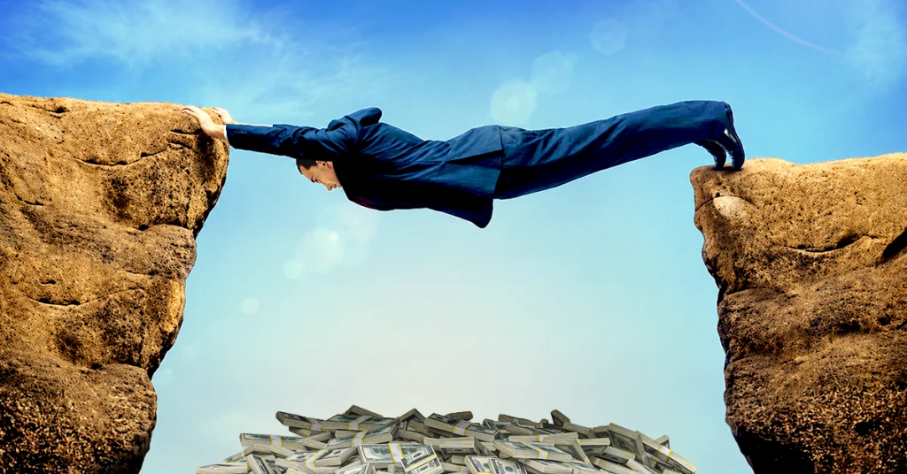 A man in a blue suit precariously holding himself up between two boulders with a pile of money beneath him.