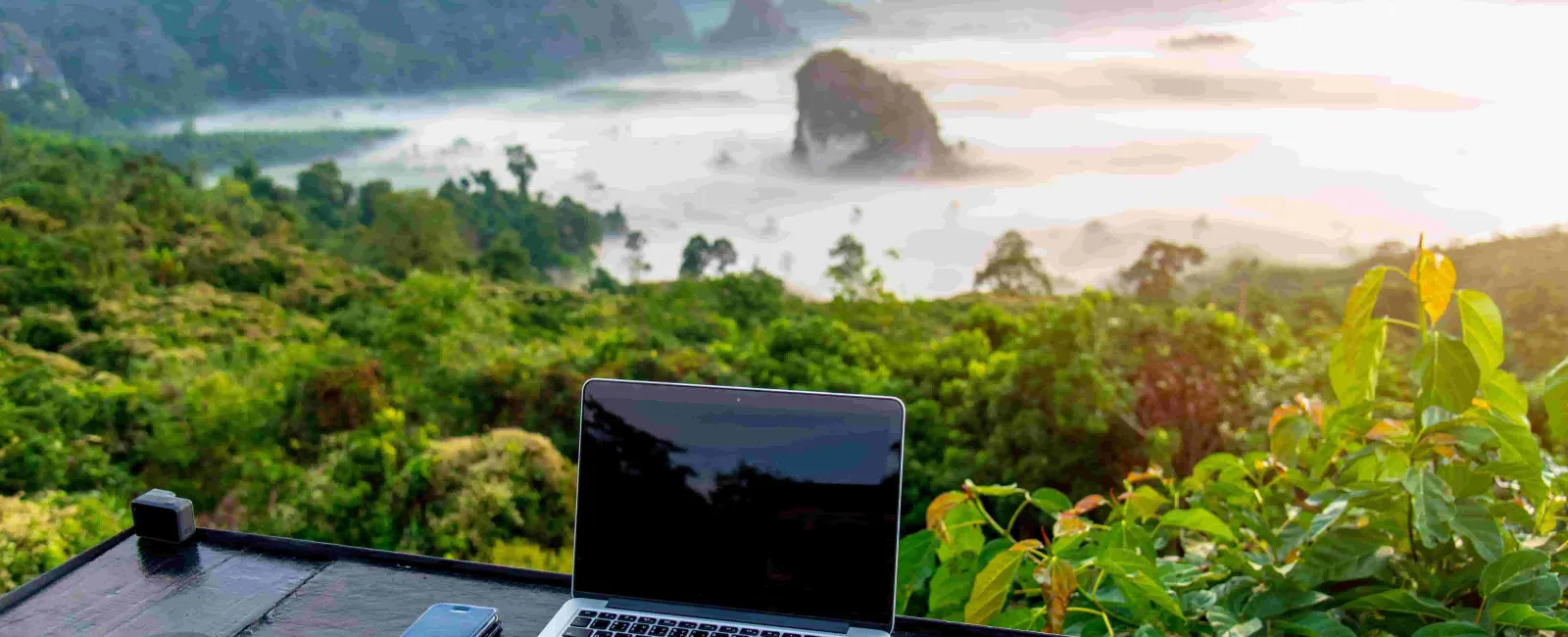 Laptop and smartphone on a wooden table overlooking green hills and misty mountains at sunrise.